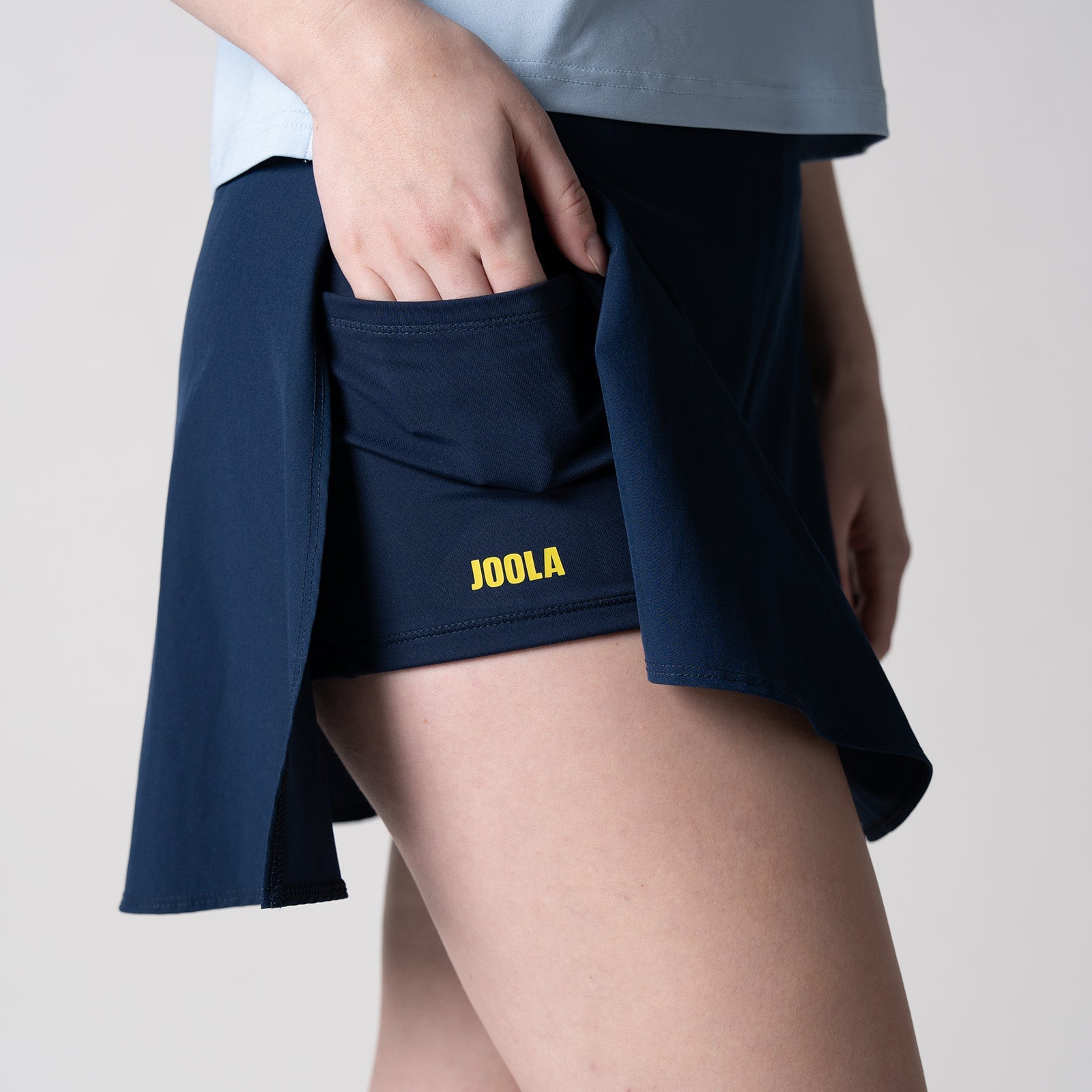 Gray background, close up image of a female model wearing the JOOLA women's pickleball skort, and revealing the hidden under-shorty pocket and yellow JOOLA word mark logo, navy.