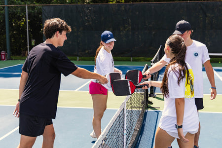 Group of four pickleball players doing a paddle tap at the net on a pickleball court.