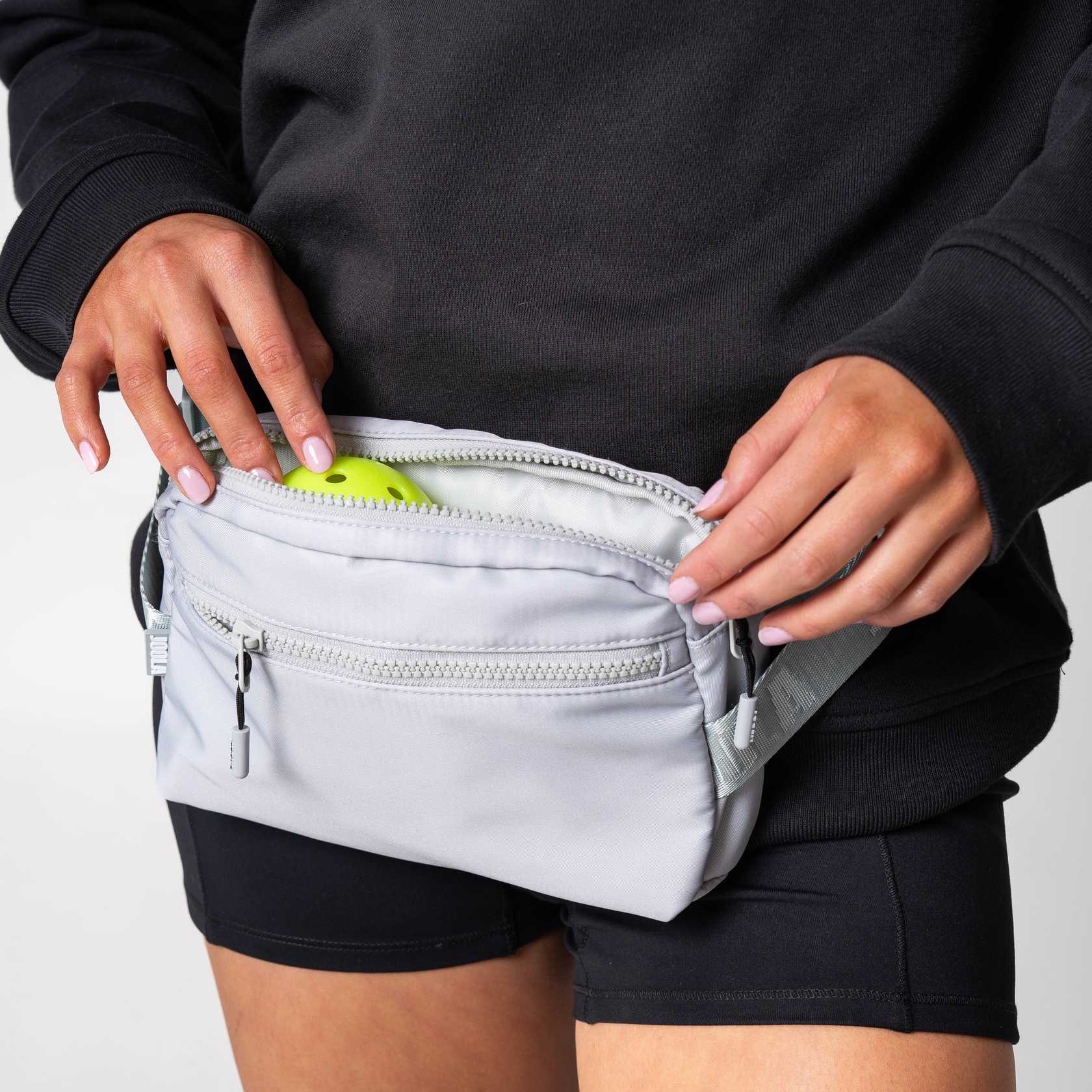 Close up image of a person putting a pickleball in to the pockey of a JOOLA Belt Bag, Gray.