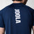 Close up on the white JOOLA brand logo that runs down the middle of the JOOLA Court Short Sleeve Shirt, Navy Blue.