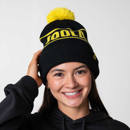 Gray background, image of a model wearing a black and yellow JOOLA Pom Pom Beanie.