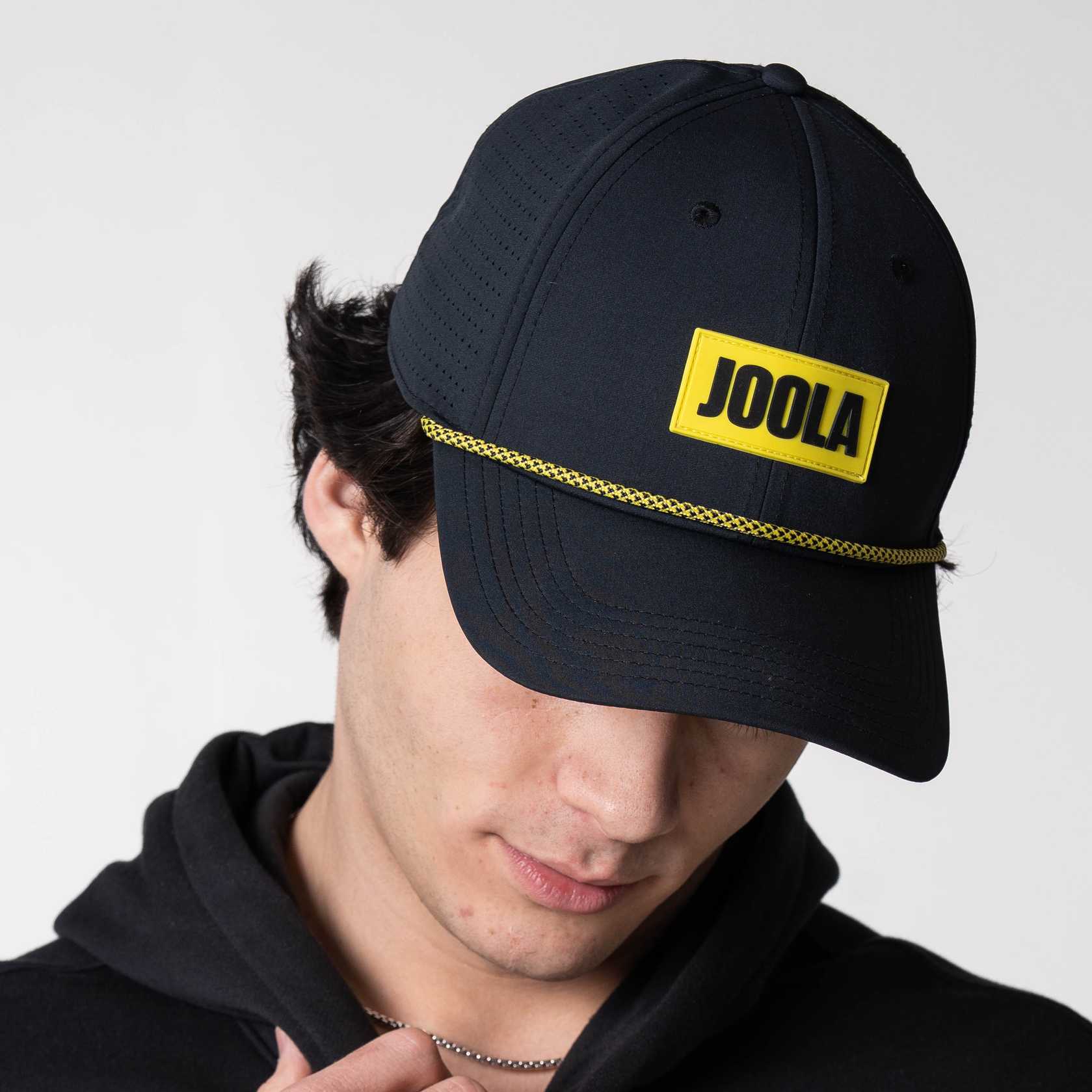Person wearing a  JOOLA Rope Cap, black with yellow branding.