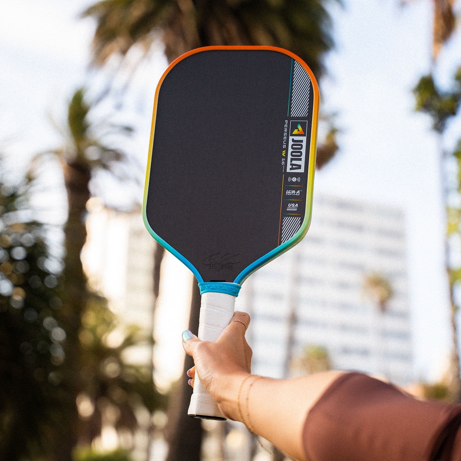 JOOLA Perseus Summer Daze Pickleball paddle with black surface, orange, blue, green and yellow color gradient on the rim, and white handle with palm trees in the background.