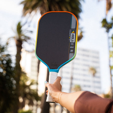 JOOLA Perseus Summer Daze Pickleball paddle with black surface, orange, blue, green and yellow color gradient on the rim, and white handle with palm trees in the background.