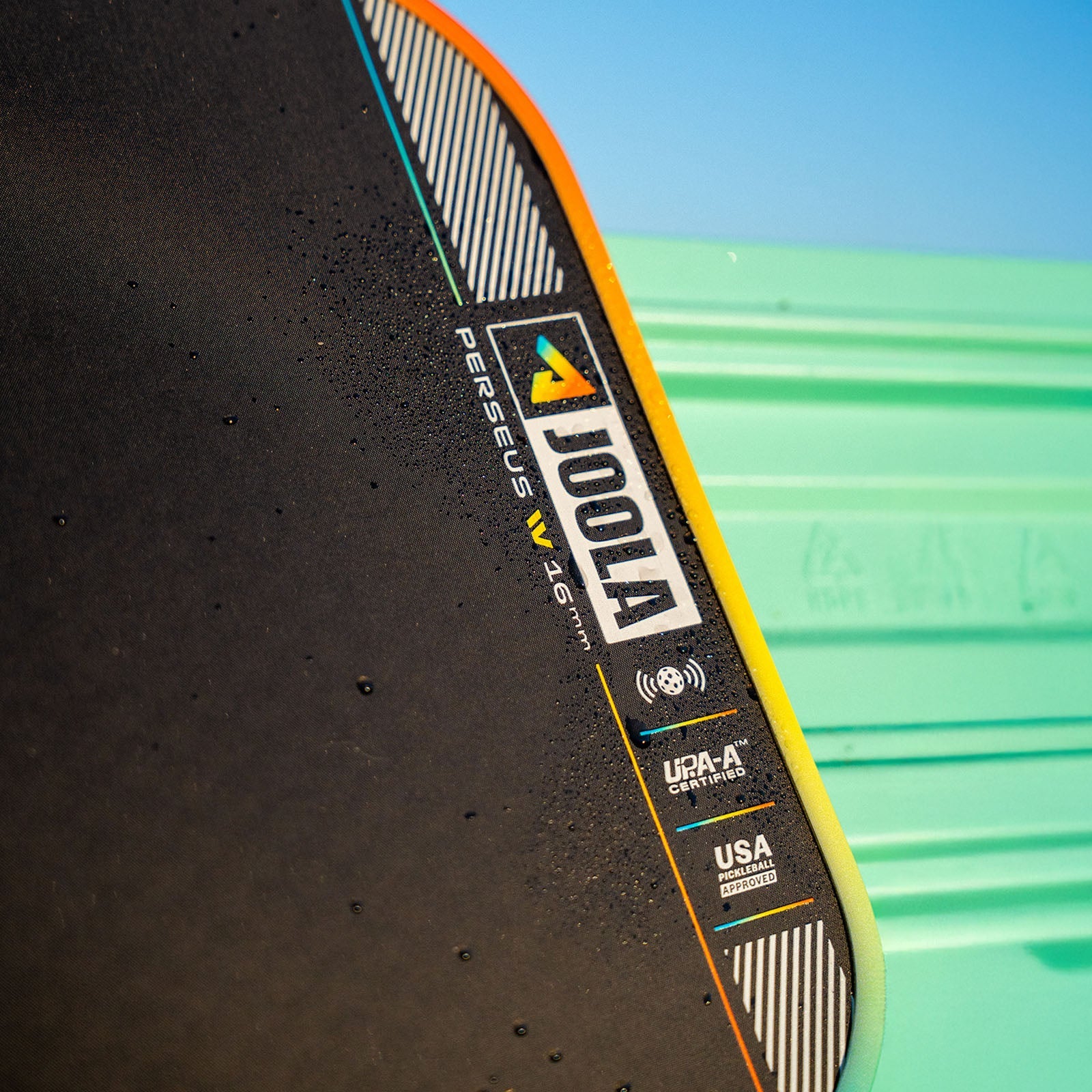 Close up image of the JOOLA Perseus Summer Daze Pickleball paddle with black surface, orange, blue, green and yellow color gradient on the rim, and white handle with a green and blue unfocused image in the background.