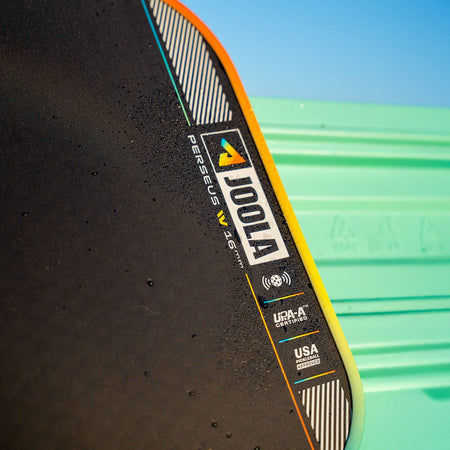 Close up image of the JOOLA Perseus Summer Daze Pickleball paddle with black surface, orange, blue, green and yellow color gradient on the rim, and white handle with a green and blue unfocused image in the background.