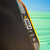 Close up image of the JOOLA Perseus Summer Daze Pickleball paddle with black surface, orange, blue, green and yellow color gradient on the rim, and white handle with a green and blue unfocused image in the background.