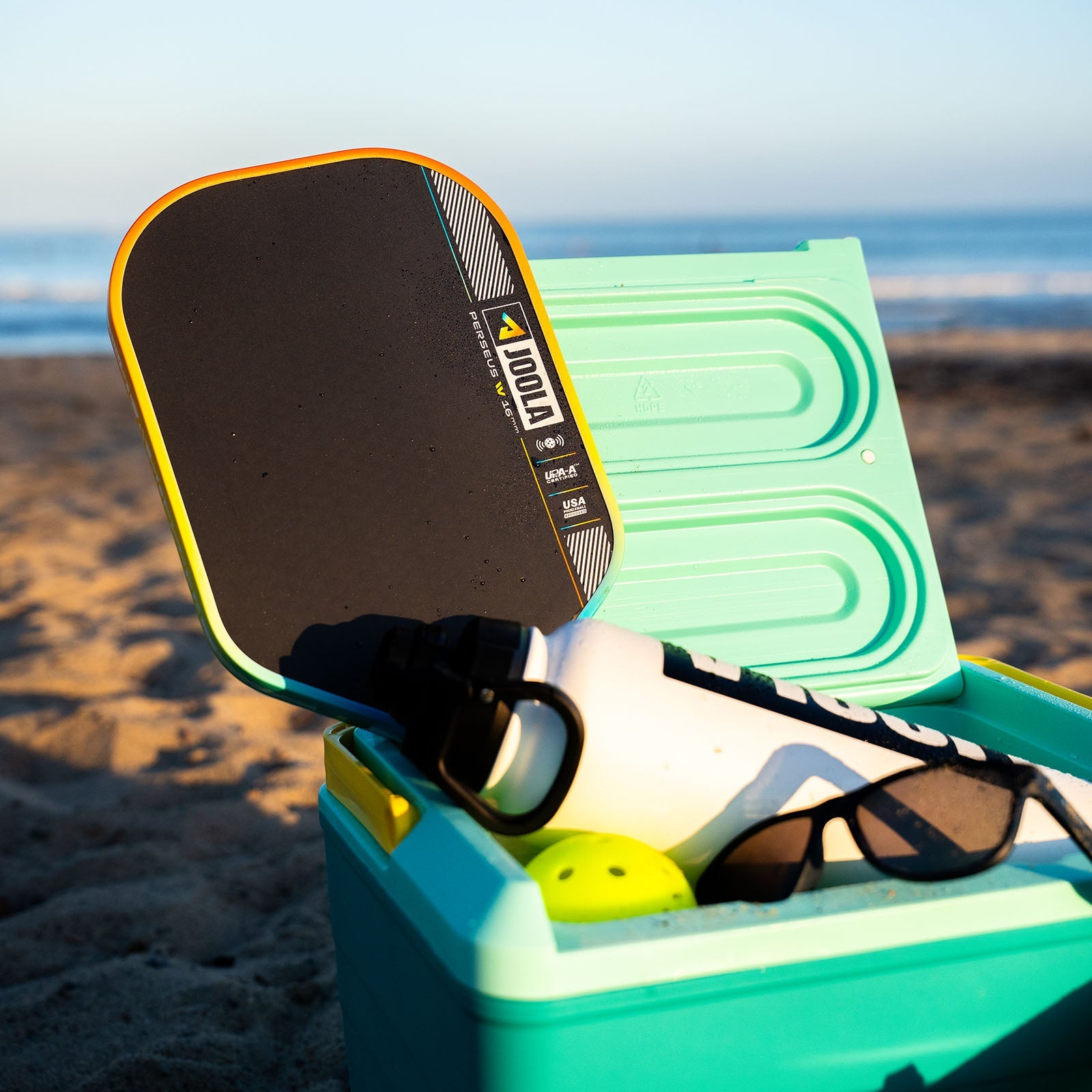 JOOLA Perseus Summer Daze Pickleball paddle with black surface, orange, blue, green and yellow color gradient on the rim, and white handle in an open cooler with items including sunglasses, and a bottle on a beach. 