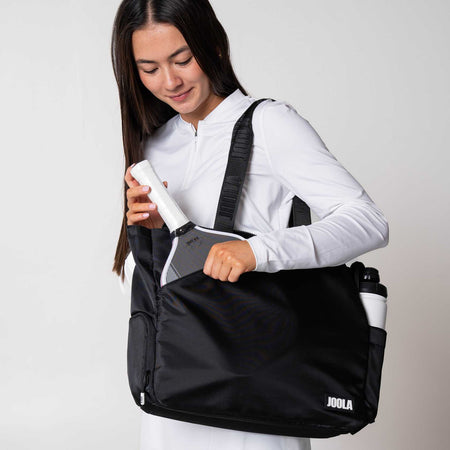 Gray background, Image of a female model putting a JOOLA Pro IV paddle in the front pocket of here JOOLA Tote bag.
